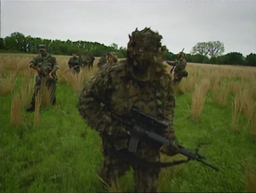 Slightly blurry still of a number of people in 1990s-style camo fatigues walking through an open field, carrying rifles