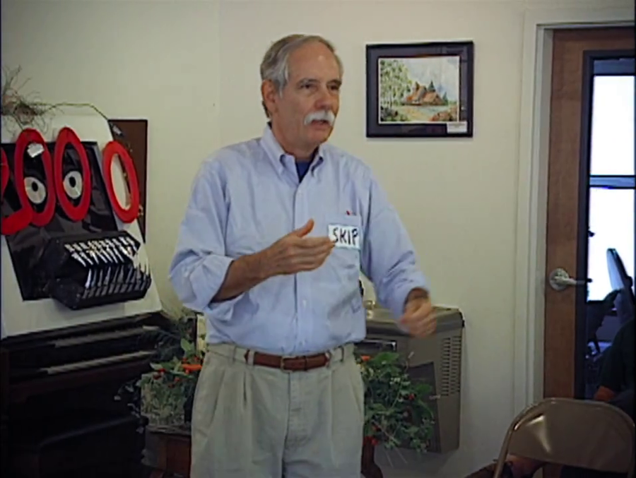 An older man with a name tag that says "Skip" and a serious expression gesticulates as he explains something. Behind him is a home-made poster featuring a monster with evil looking eyes and many teeth made of paper protruding from the poster, as well the digits "2000" over the monster's face.