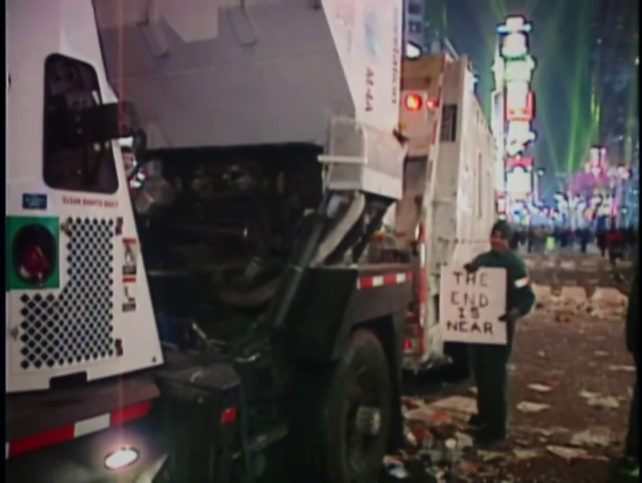 A sanitation worker stands on trash-strewn ground, next to some garbage trucks, showing a paperboard sign to the camera and grinning